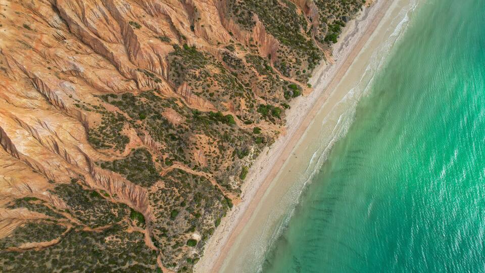 A coastal landscape with a steep cliff, a sandy beach, and turquoise waters.