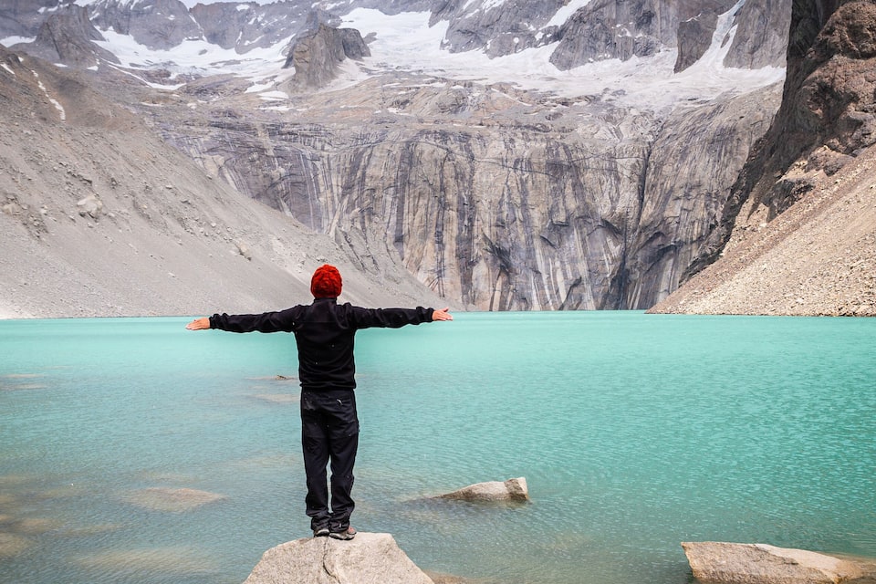 A person standing on a rock with arms outstretched facing a mountain range and a turquoise lake.