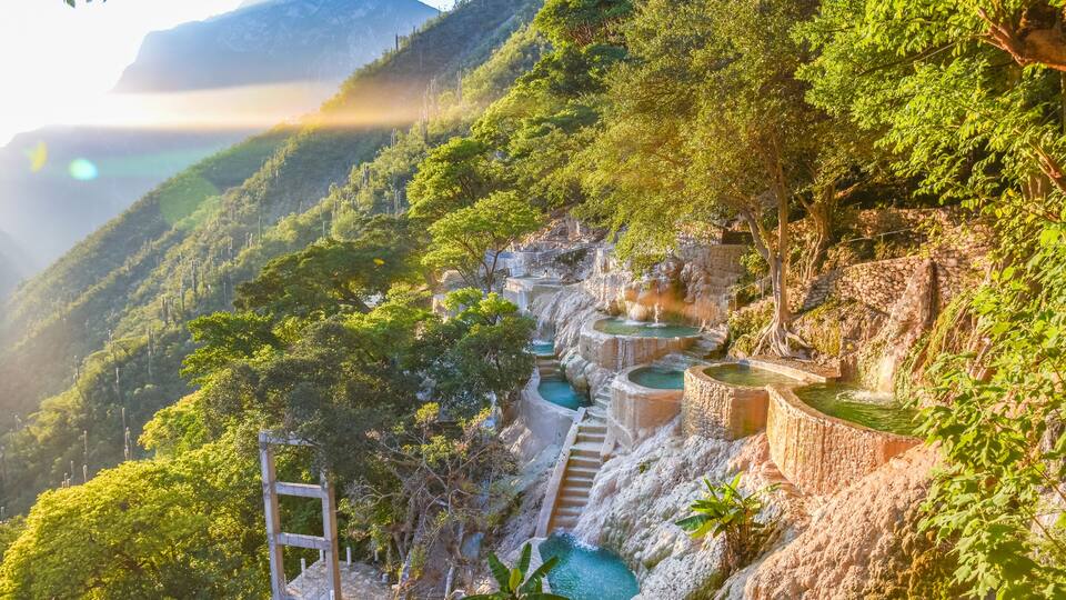 Terraced pools on a hillside, surrounded by lush greenery and mountains in the background.