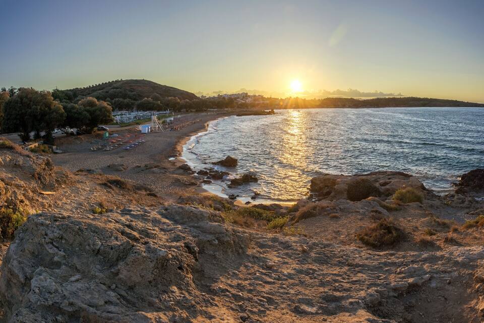 A beach with the sun setting over the water, rocky terrain, and a distant hill with buildings.