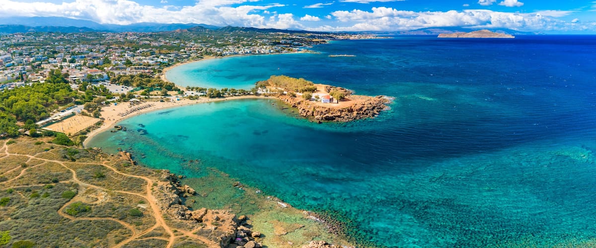 An aerial view of a coastal town with clear turquoise waters, a rocky island, and a distant mountain range.