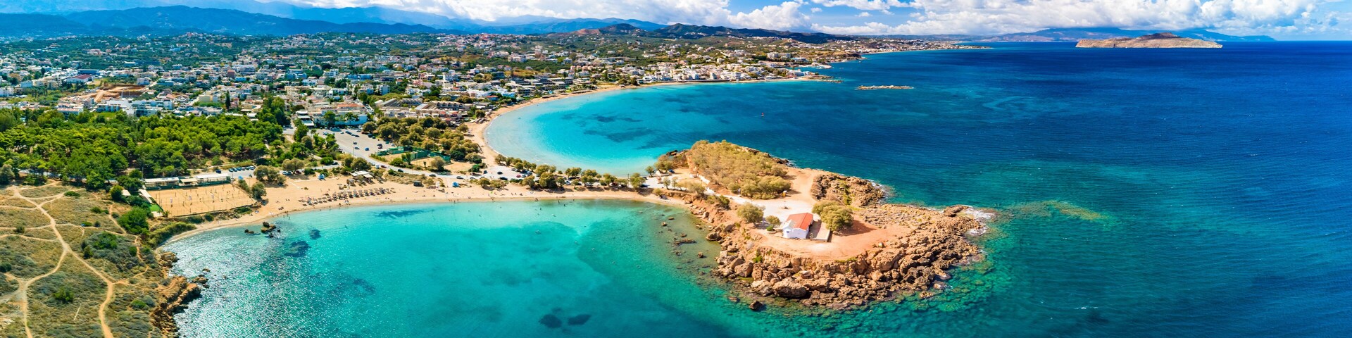 Vista aérea de un pueblo costero con una playa, aguas turquesas cristalinas y un promontorio rocoso.