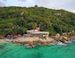 A coastal area with a building on a rocky outcrop, surrounded by lush green hills and clear turquoise waters.
