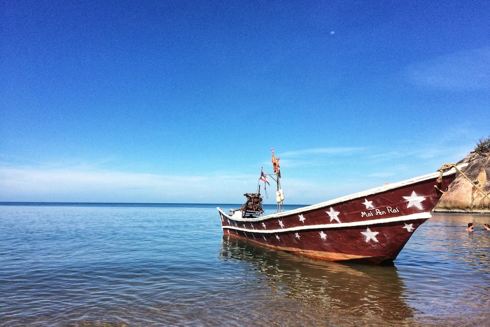 Ein Boot namens "Mai Pen Rai" ankert in der Nähe einer felsigen Küste mit klarem, blauem Wasser.