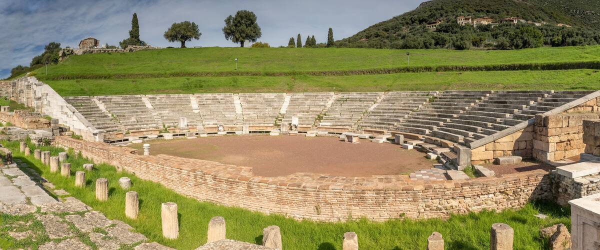 An ancient amphitheater with stone steps and a central stage, surrounded by grassy hills and trees.