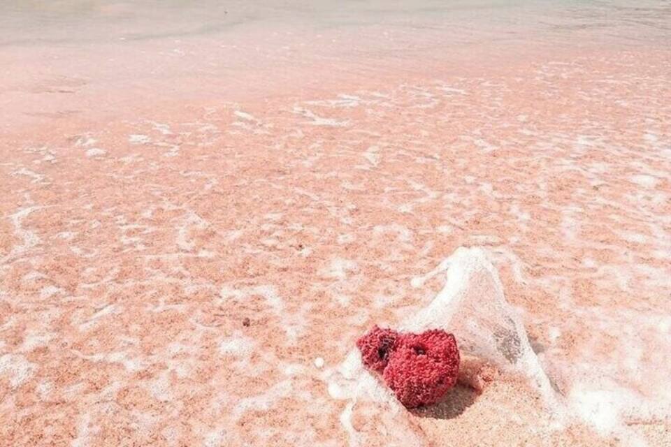 A pink sandy beach with a heart-shaped object in the foreground and boats in the background.