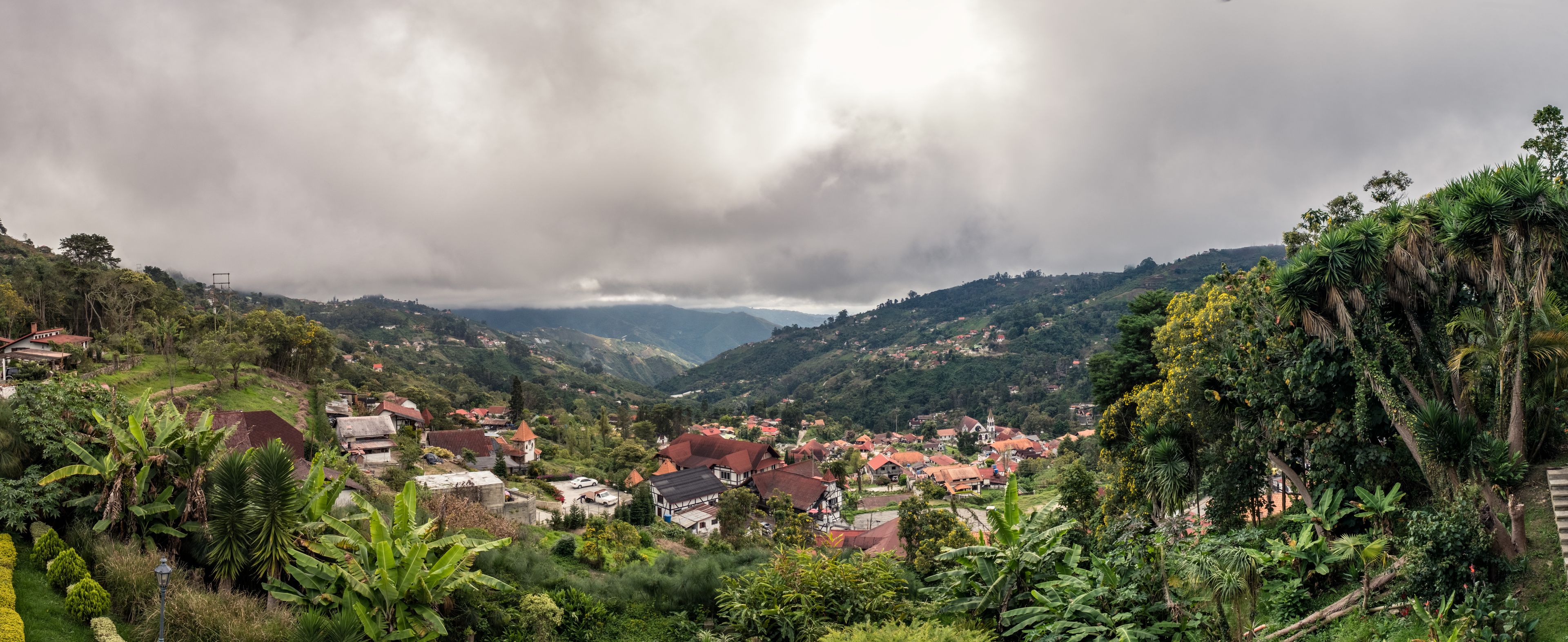 Une vallée verdoyante et luxuriante, parsemée de maisons aux toits rouges, d’une végétation dense et surplombée d’un ciel nuageux.