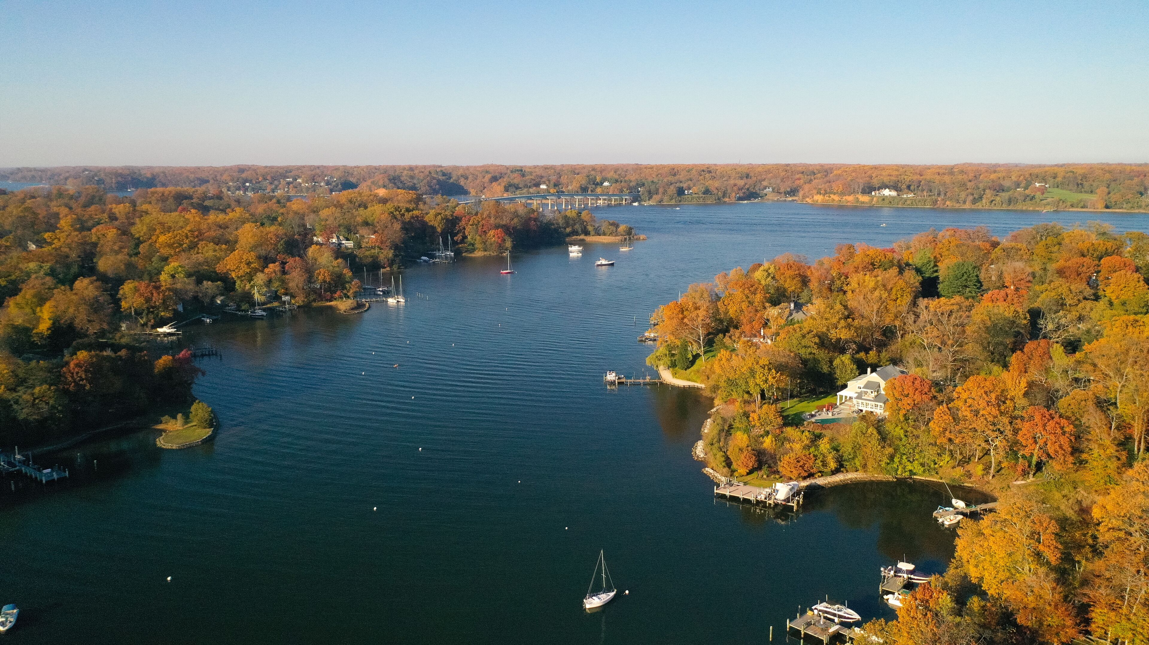A river with boats, surrounded by autumn-colored trees and a bridge in the distance.