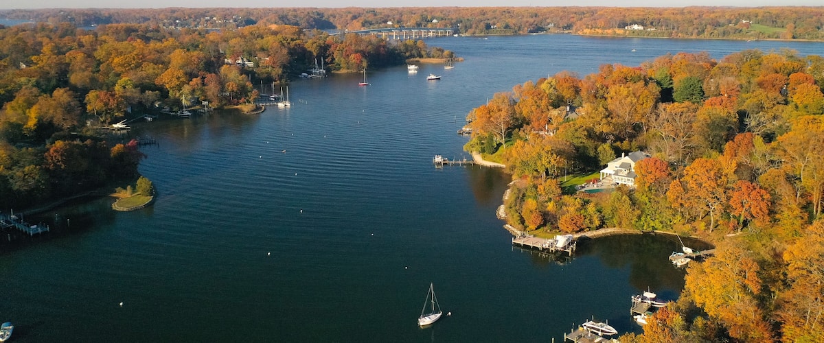 A river with boats, surrounded by autumn-colored trees and a bridge in the distance.