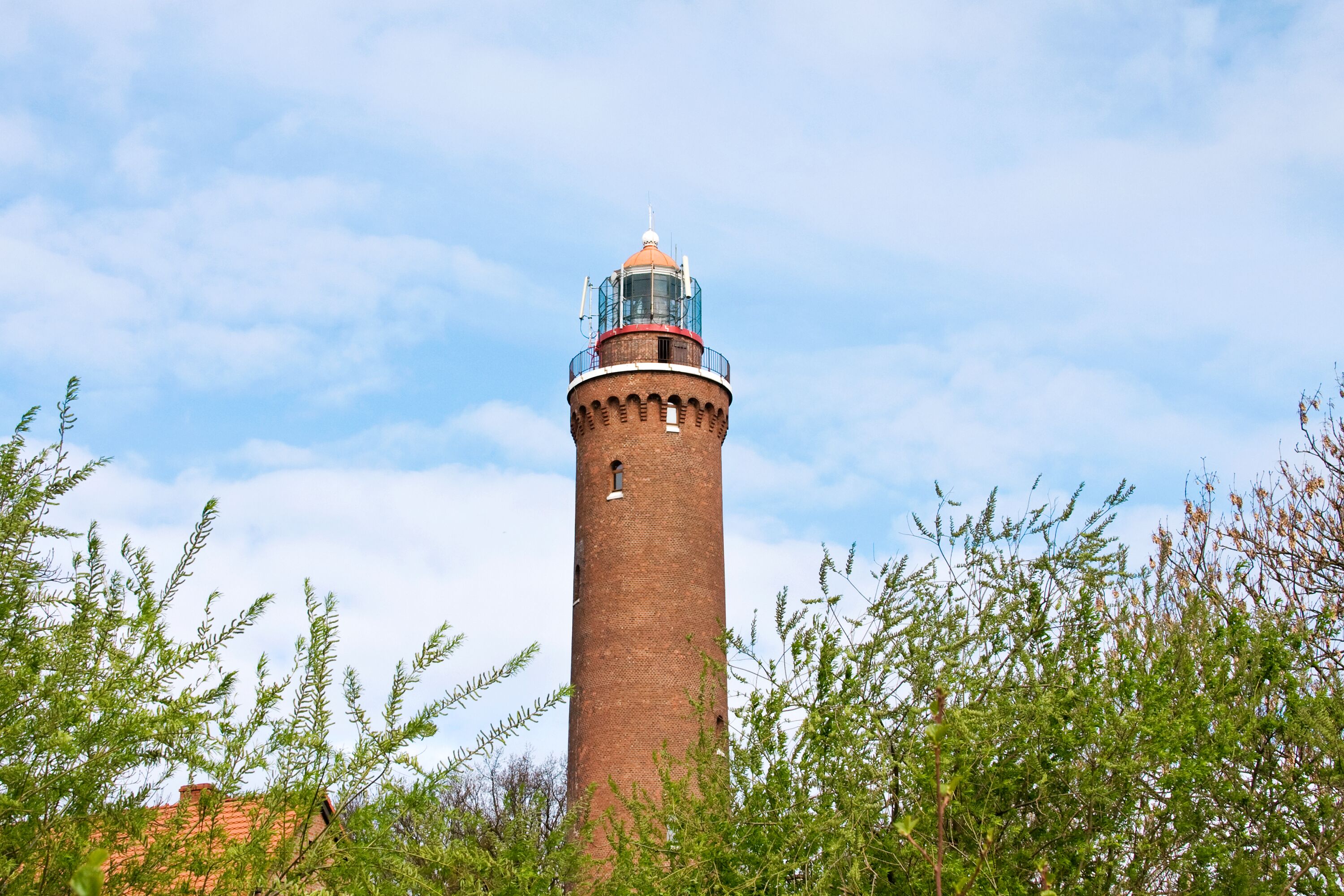 A tall brick lighthouse with a red roof and a white dome, surrounded by green foliage.