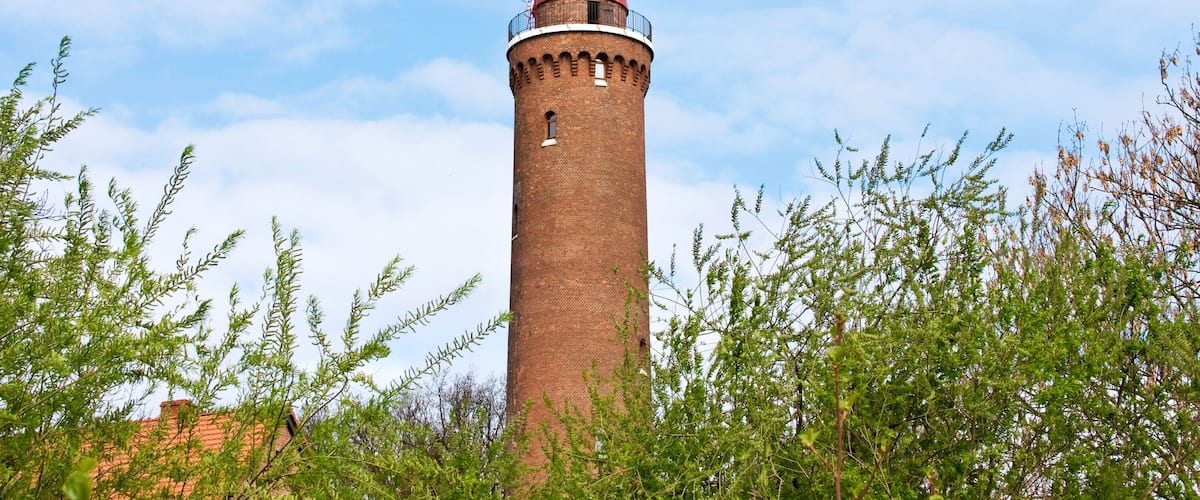 A tall brick lighthouse with a red roof and a white dome, surrounded by green foliage.