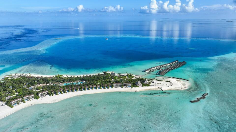 An aerial view of a tropical resort with clear turquoise waters, a sandy beach, and a long pier extending into the sea.