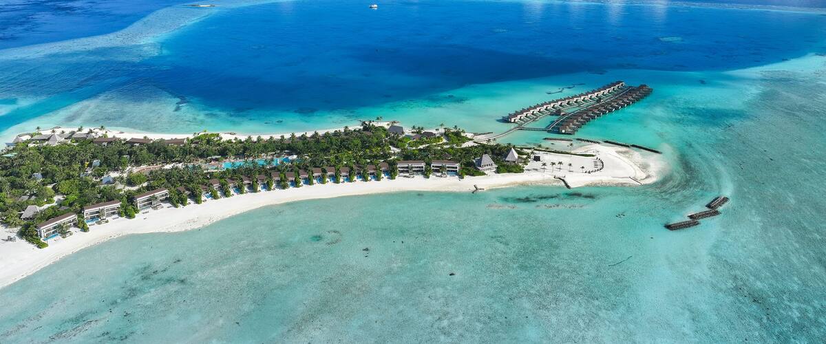 An aerial view of a tropical resort with clear turquoise waters, a sandy beach, and a long pier extending into the sea.