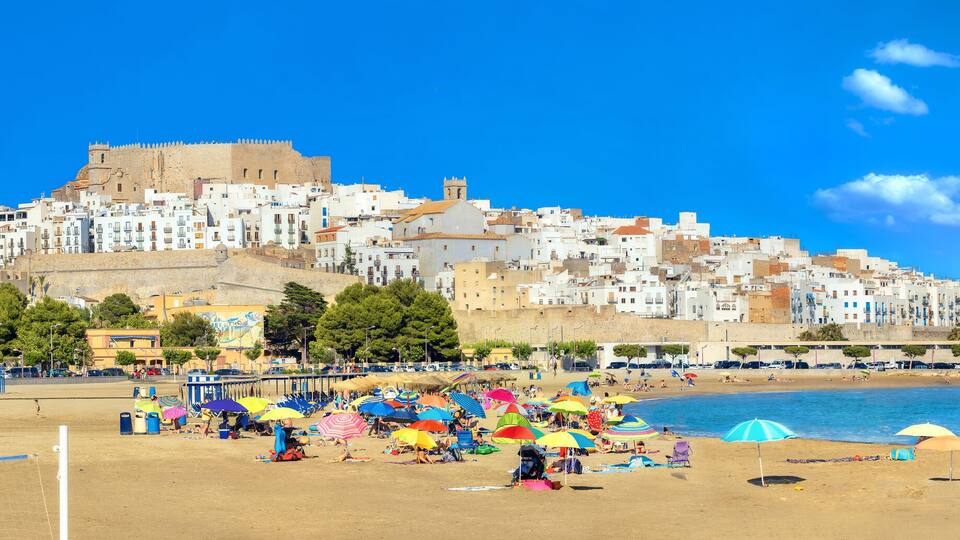 En strand med färgglada parasoller och folk som solar, en sandstrand och en historisk kulle med byggnader i bakgrunden.