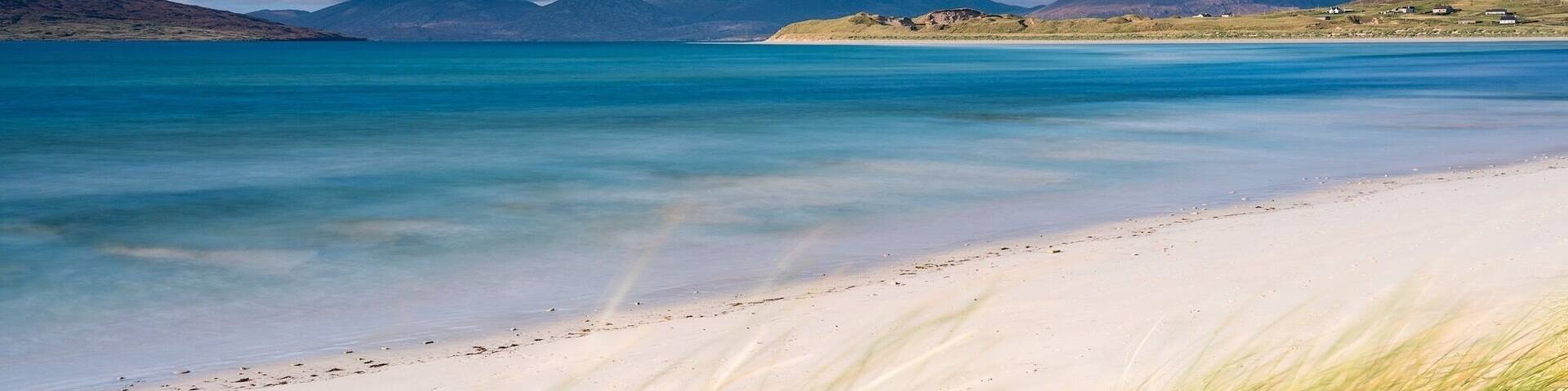 Una playa con agua turquesa cristalina, arena blanca y dunas cubiertas de hierba.