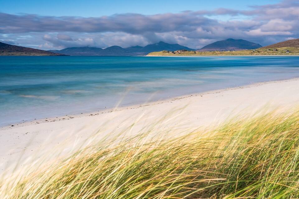 A beach with clear turquoise water, white sand, and grassy dunes.