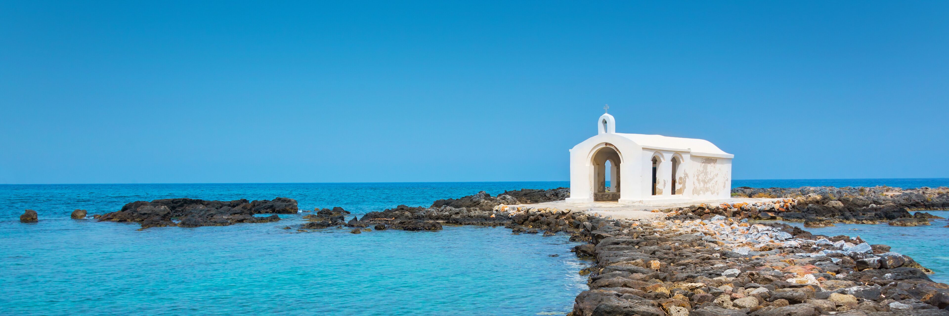 A white church on a rocky shoreline with clear blue water.