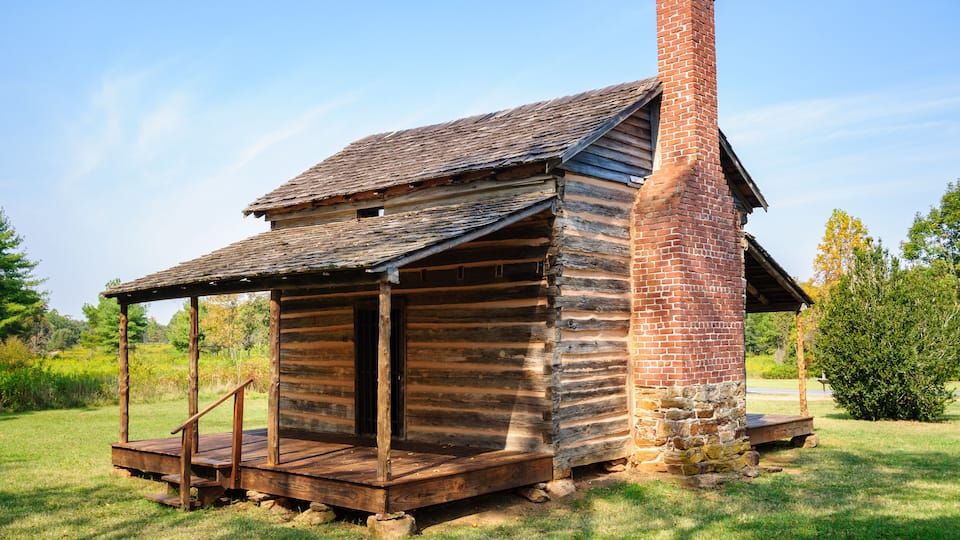 A rustic log cabin with a brick chimney, surrounded by lush greenery.