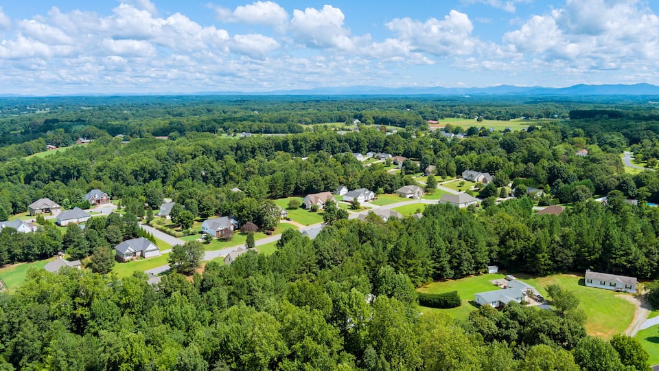 A residential area with houses surrounded by dense forest and rolling hills in the background.