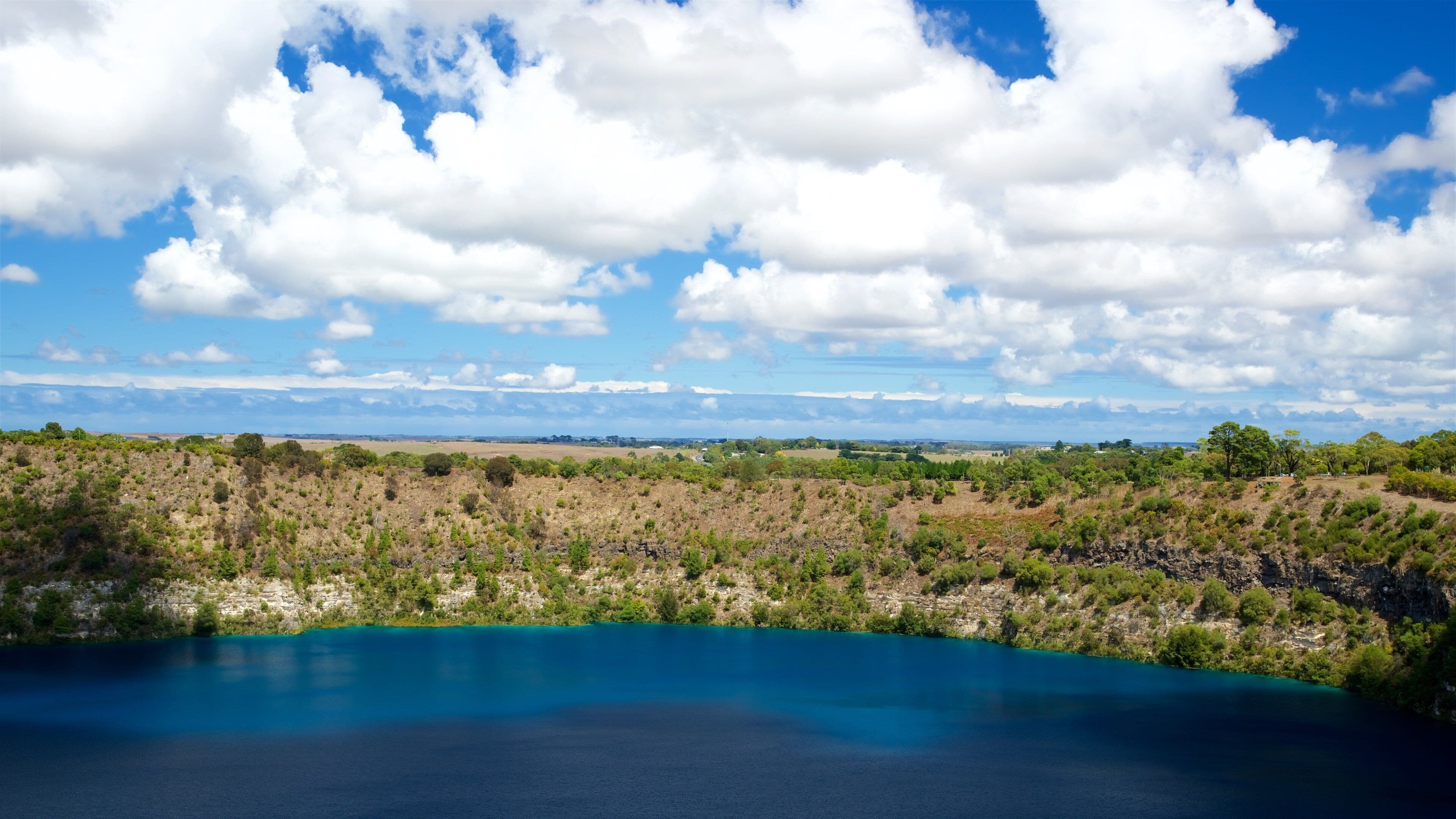 A coastal landscape with a clear blue sea, green vegetation, and a distant view of the horizon under a partly cloudy sky.