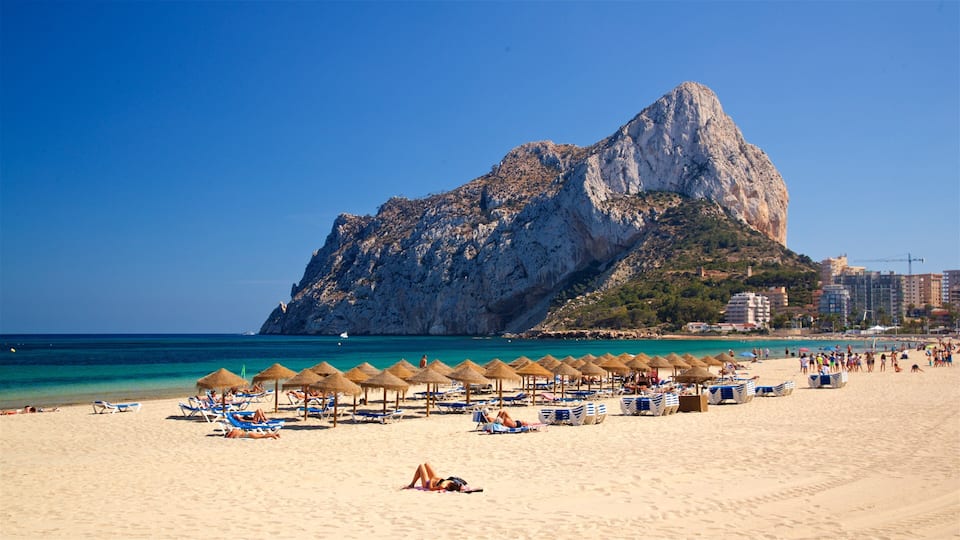 A beach with thatched umbrellas, sun loungers, and a large rocky mountain in the background.