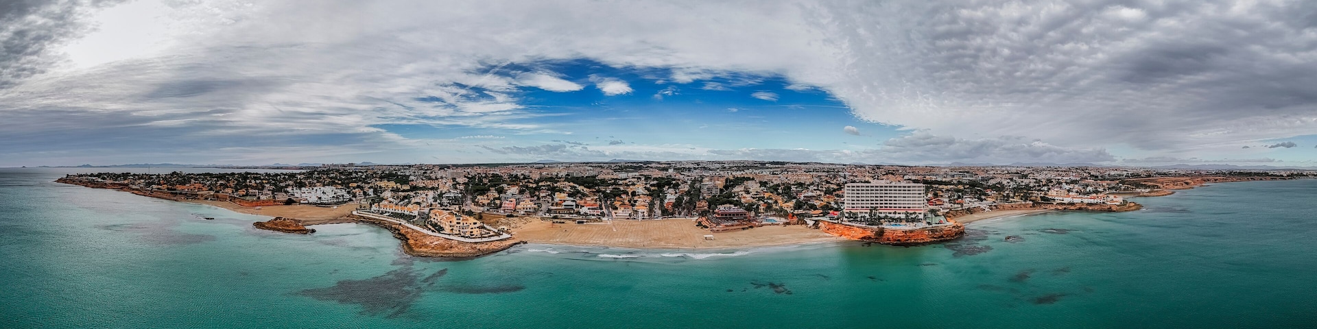 An aerial view of a coastal city with a prominent beach, a large hotel, and a mix of residential and commercial buildings.