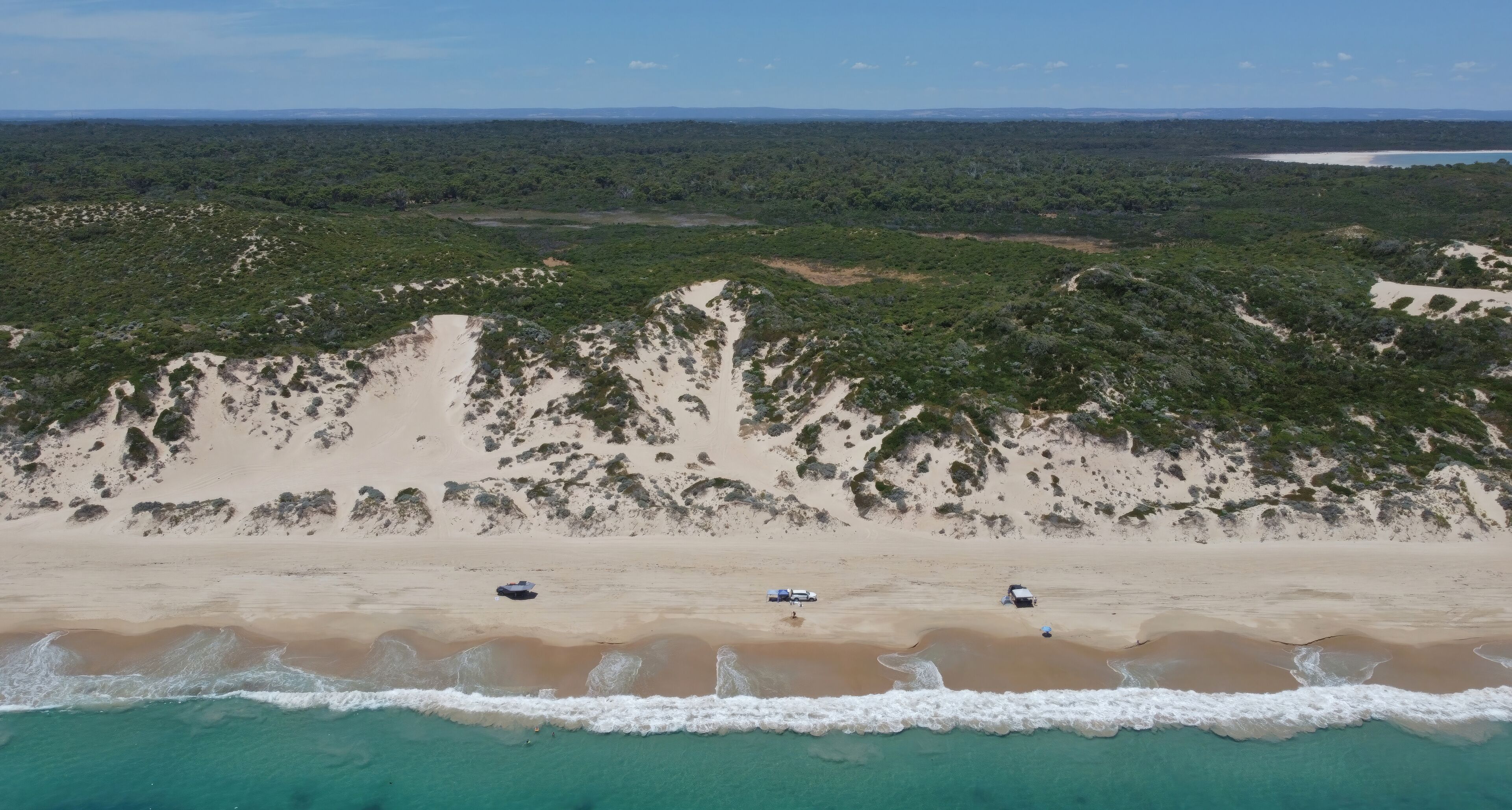A beach with clear turquoise waters, sandy dunes, and sparse vegetation.