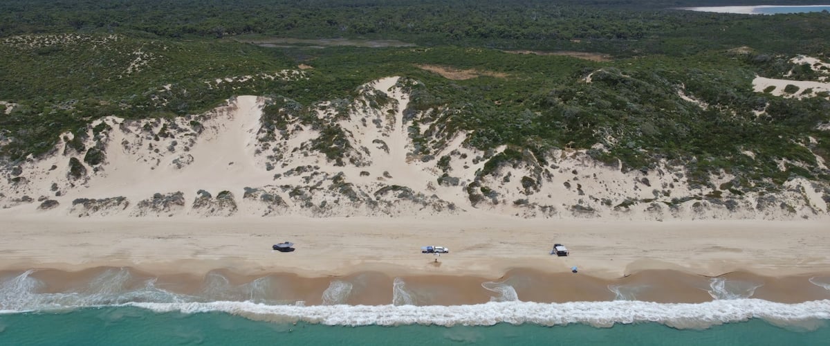 A beach with clear turquoise waters, sandy dunes, and sparse vegetation.