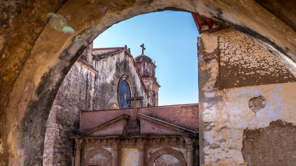 An arched doorway framing a weathered church with a cross on top and a large circular window.