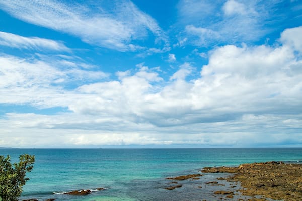 Un paysage côtier avec une eau bleue et limpide, une rive rocheuse et un ciel partiellement nuageux.