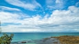 A coastal landscape with clear blue water, rocky shore, and a partly cloudy sky.