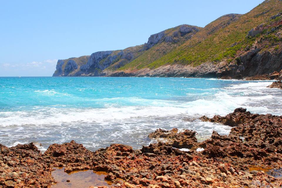 A rocky coastline with clear blue waters and a mountainous backdrop.