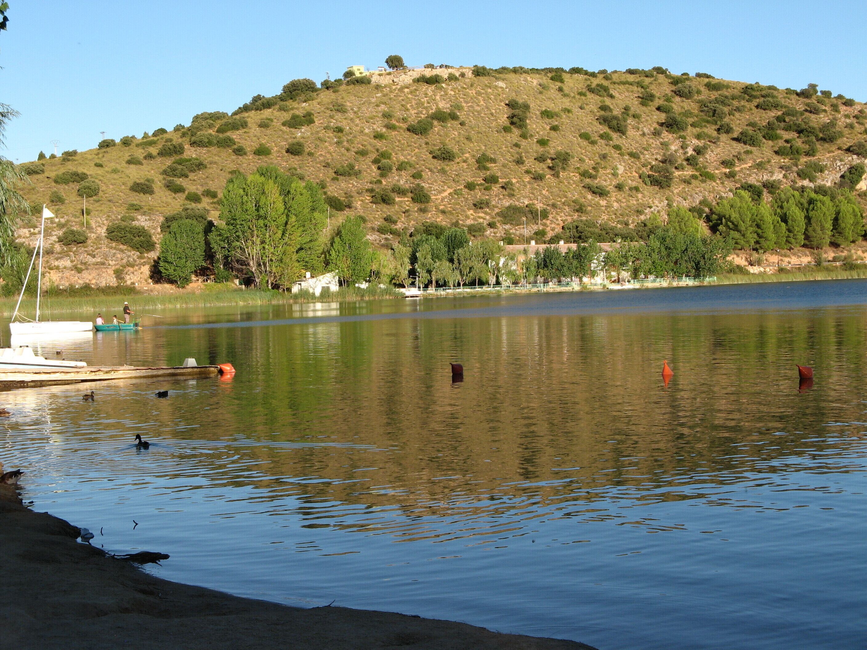 Um lago tranquilo com barcos, uma margem arenosa e uma colina com árvores ao fundo.