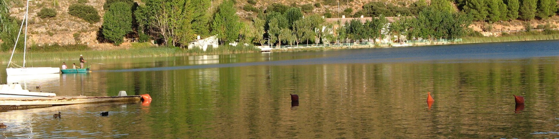 Um lago tranquilo com barcos, uma margem arenosa e uma colina com árvores ao fundo.