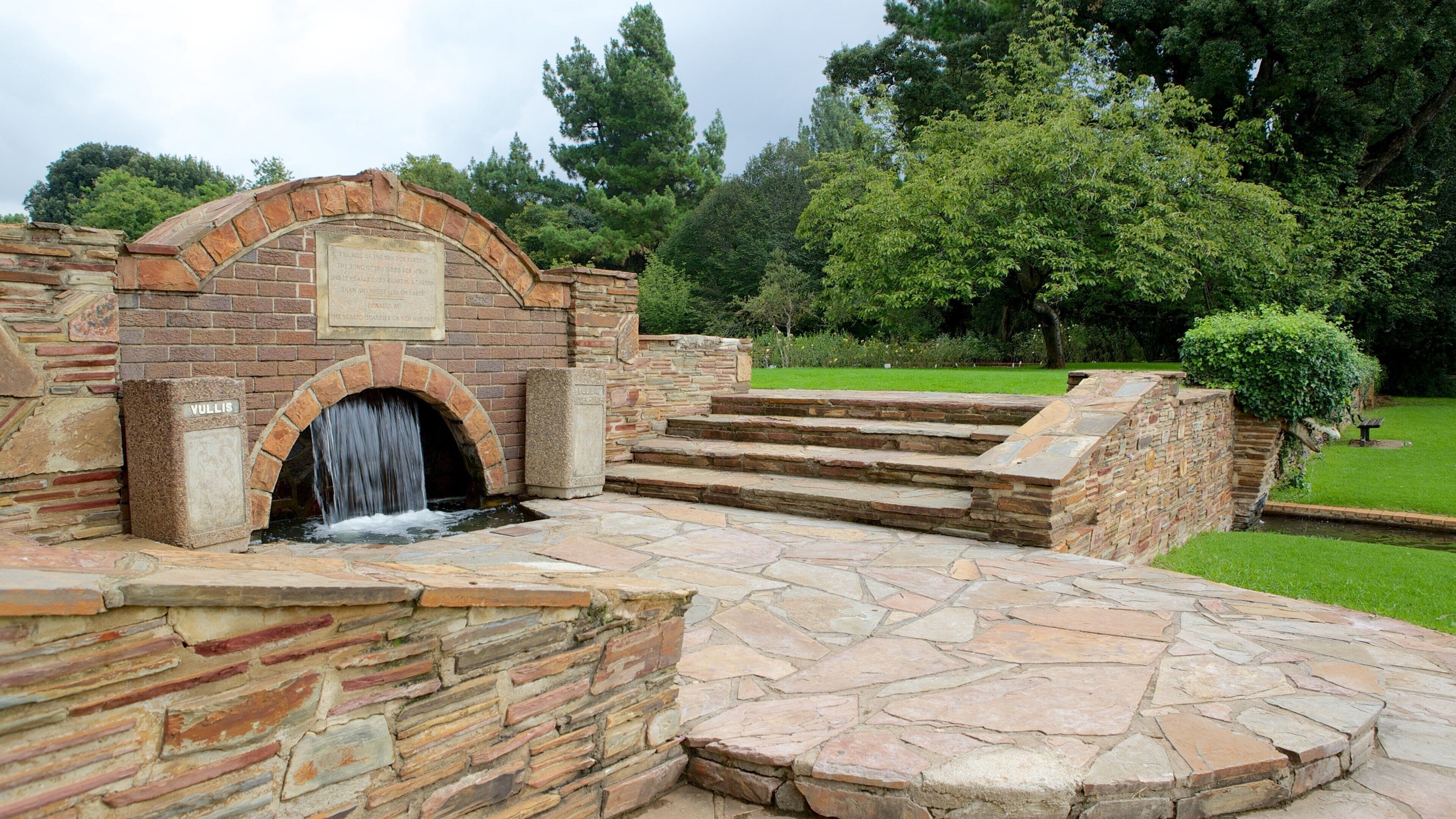 Une arche en pierre surmontée d’une cascade, avec des marches menant à celle-ci et une plaque commémorative.