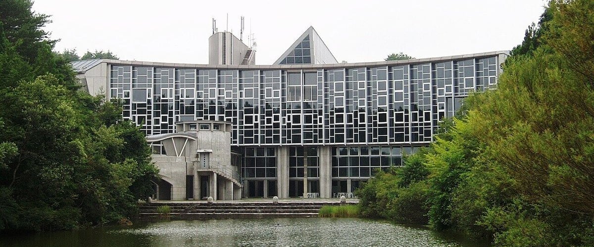 A modern building with a geometric facade, surrounded by a body of water and lush greenery.