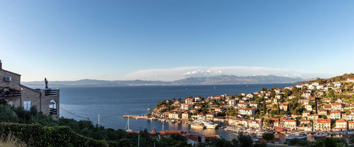 A coastal town with buildings on a hillside, a marina with boats, and a clear view of the sea and distant mountains.