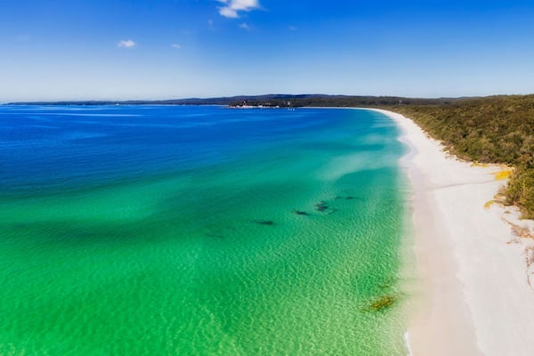 A coastal landscape with a white sandy beach, clear turquoise waters, and a dense forested area.