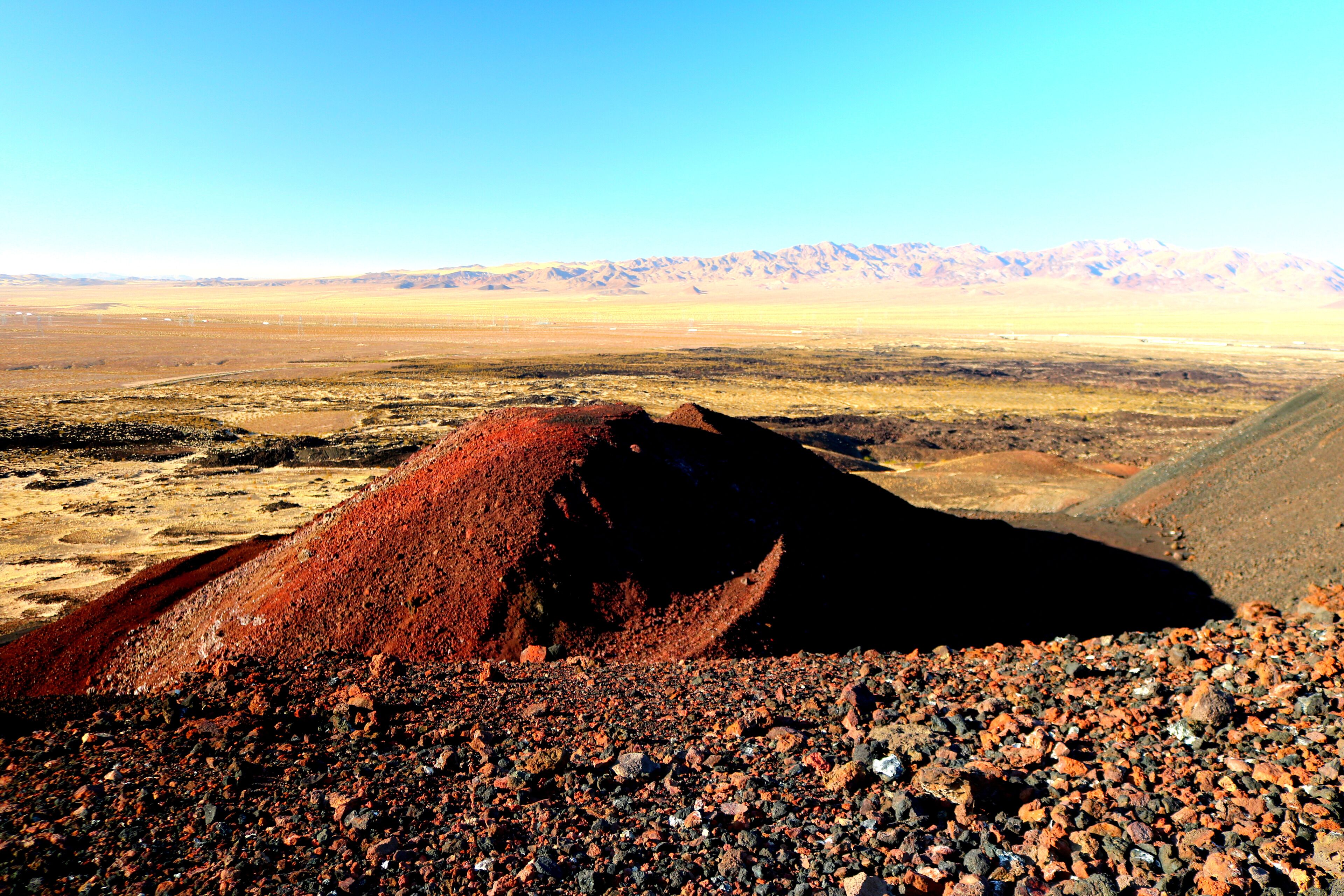A barren landscape with a large pile of red earth in the foreground and a mountain range in the background.