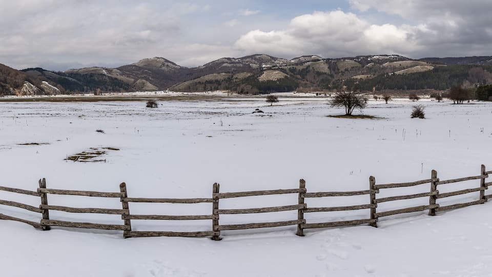 A snow-covered field with a wooden fence and mountains in the background.