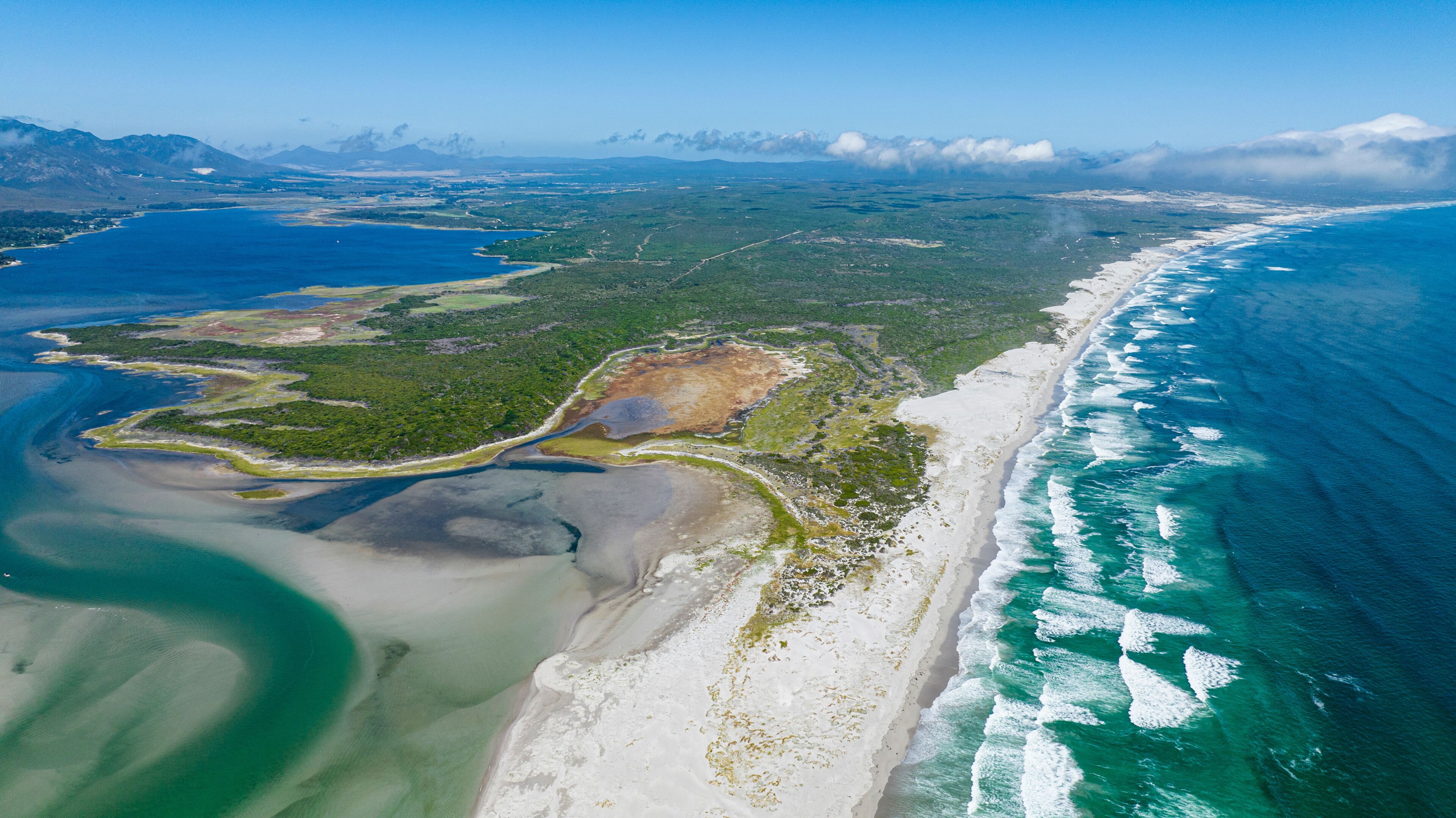 An aerial view of a coastal area with a sandy beach, a clear blue ocean, and a green landscape with a winding road.