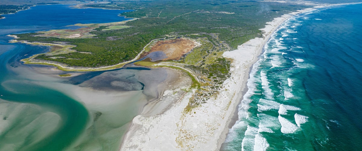 An aerial view of a coastal area with a sandy beach, a clear blue ocean, and a green landscape with a winding road.