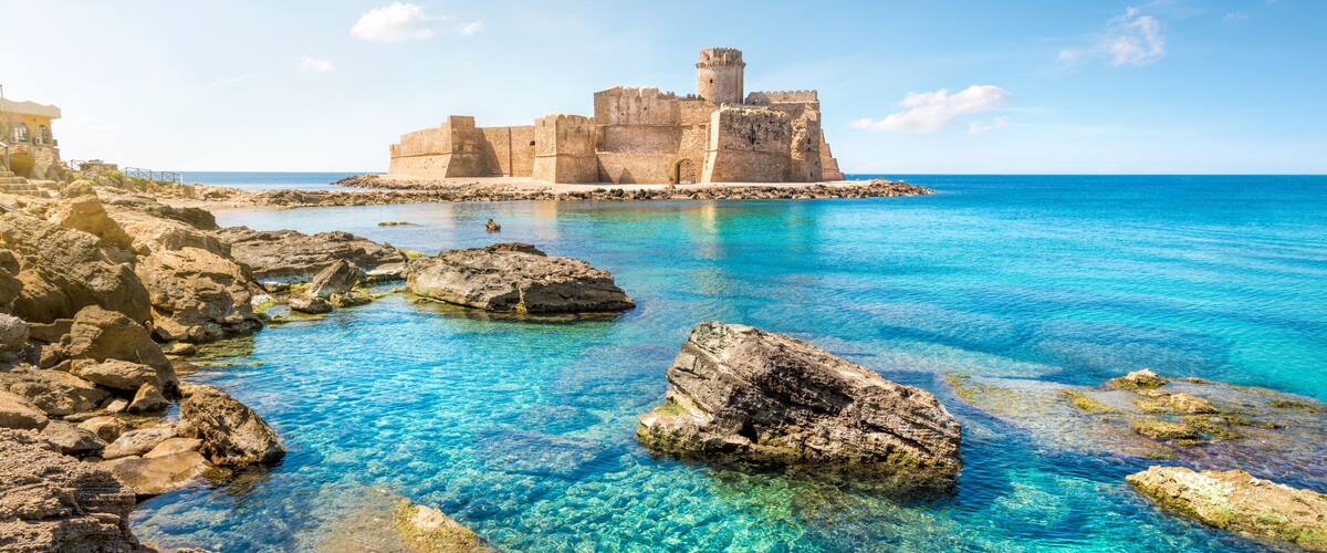 A coastal castle surrounded by clear blue waters and rocky shores.