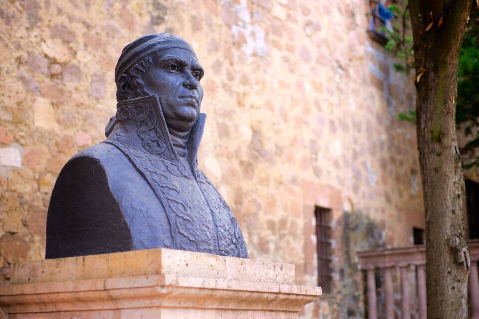 A statue of a historical figure in a stone building with a tree and a railing in the background.