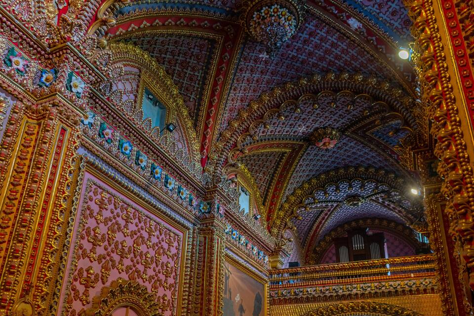 An ornate church interior with intricate golden decorations, a large mural, and a high vaulted ceiling.