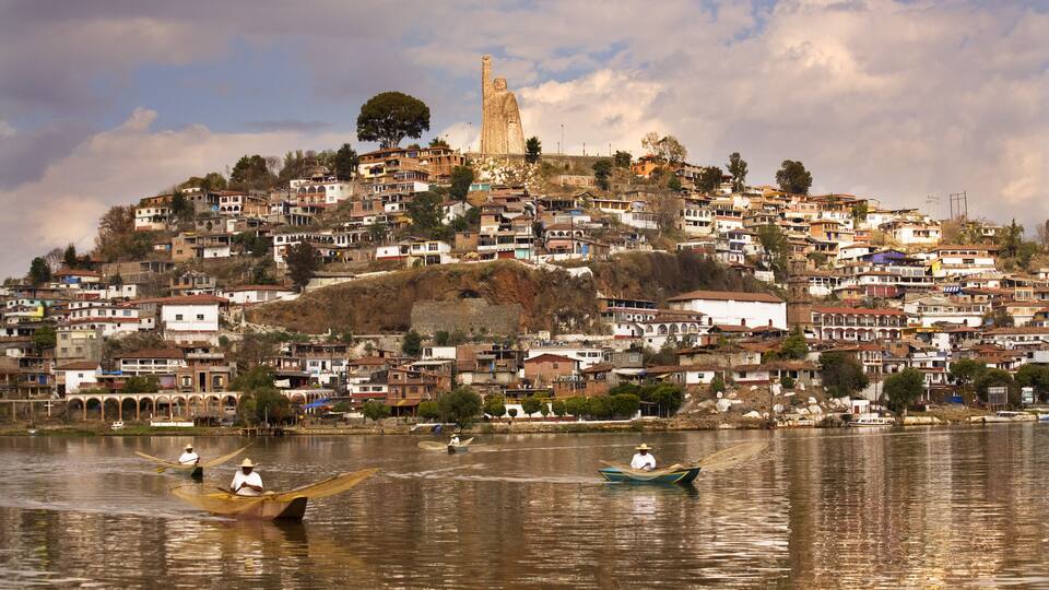 A hillside town with a prominent statue on top, boats on a calm river, and dense housing.