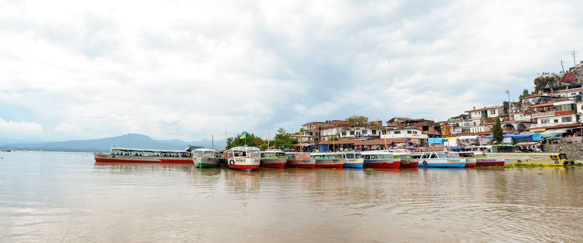A harbor with several boats docked along the shore, surrounded by a hillside with buildings and a mountain in the background.