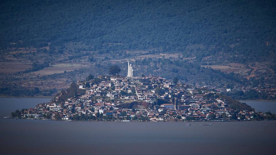 A coastal town with a prominent statue on a hill, surrounded by dense housing and a large body of water.