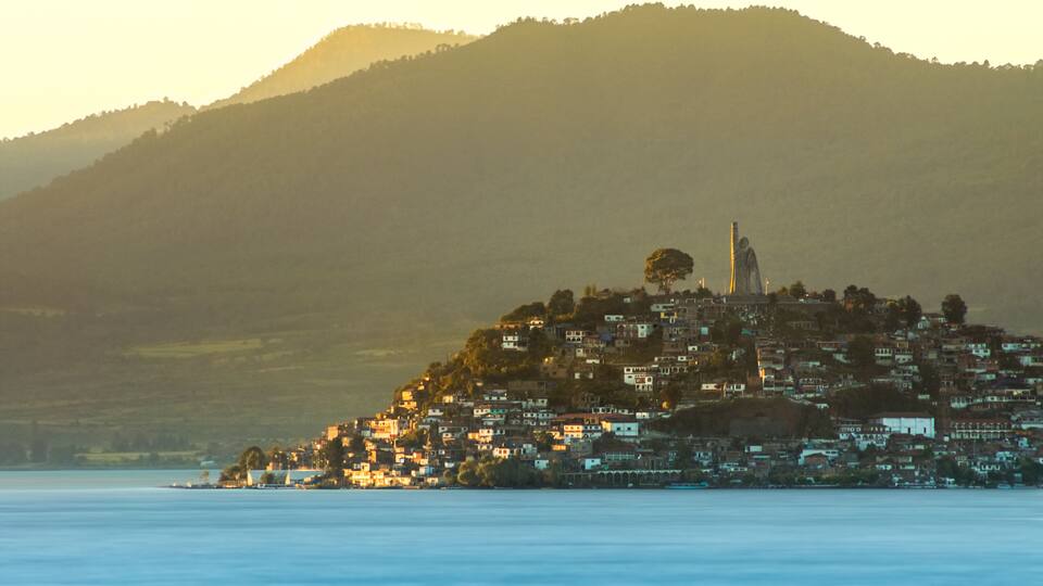 A coastal town with a prominent statue on a hill, surrounded by houses and a mountain in the background.