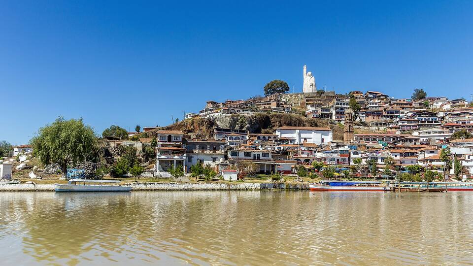 A coastal town with a prominent statue on a hill, houses along the waterfront, and a body of water reflecting the buildings.